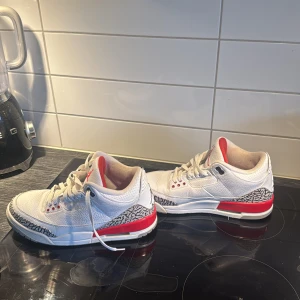 Nike Air Jordan 3 Retro hall of fame - Säljer ett par vita Nike Air Jordan 3 Retro med röda detaljer och klassiskt elefantmönster vid tån och hälen. Skorna har snörning, rund tå och ikonisk Jumpman-logga på baksidan. Perfekta för dig som gillar streetwear och sneakerskultur. Finns kvitto