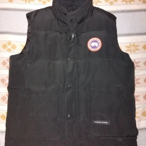 Svart dunväst från Canada Goose - Snygg svart dunväst från Canada Goose med broderad logotyp på bröstet. Västen har både dragkedja och knappar framtill för extra skydd mot kyla. Perfekt för kyliga dagar.