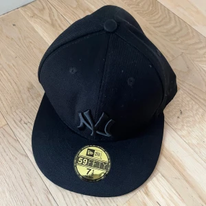 Svart New Era NY keps 59FIFTY - Säljer en svart New Era 59FIFTY keps med NY-logga framtill. Klassisk modell med rak skärm och broderad logga i svart ton-i-ton. Passar dig som gillar stilrena accessoarer och streetwear.