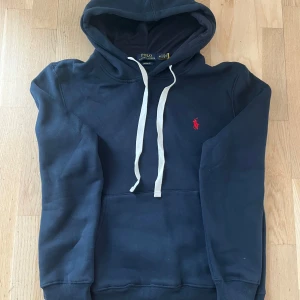 Mörkblå hoodie från Polo Ralph Lauren - Säljer en mörkblå hoodie från Polo Ralph Lauren med vit dragsko och klassisk röd logga broderad på bröstet. Tröjan har känguruficka och mjuk bomullskänsla. Perfekt för en avslappnad stil.
