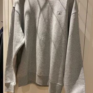 Grå sweatshirt från Champion - Säljer en klassisk grå sweatshirt från Champion i modellen Oxford Gray. Tröjan har rund hals, långa ärmar och diskret Champion-logga broderad på bröstet. Aldrig använd då den var för stor. Det står M i den men skulle säga att den passar L bättre.