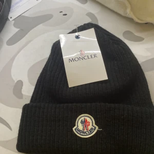 Svart moncler mössa - Svart ribbstickad mössa från Moncler med uppvikt kant och klassisk Moncler-logga framtill. Perfekt accessoar för kyliga dagar och snygg detalj till din outfit.