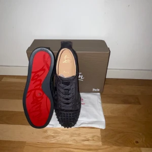 Gråa sneakers med nitar från Christian Louboutin - Säljer ett par gråa sneakers från Christian Louboutin med ikonisk röd sula och gråa nitar framtill. Skorna har snörning och är tillverkade i skinn med en matt finish. Perfekta för dig som vill sticka ut med en lyxig och edgy stil.
