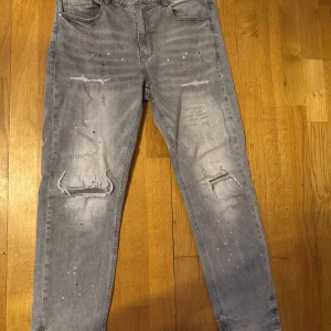 Grå jeans med slitningar från Supply & Demand - Säljer ett par grå jeans från Supply & Demand med slitna detaljer och färgstänk på framsidan. Byxorna har fem fickor, normal passform och klassisk knappgylf. Perfekta för en avslappnad streetstil.