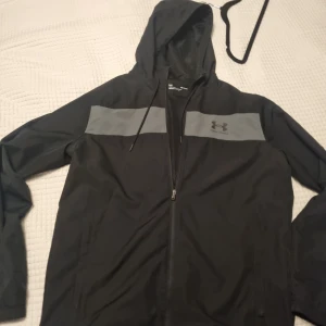 Under Armour jacka - Under Armour jacka med gråa detaljer över bröstet och logga framtill. Jackan har huva med dragsko, dragkedja och långa ärmar. Sparsamt använd, ser ut som ny.