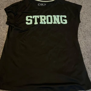 Svart t-shirt med STRONG-tryck från OTP - Svart t-shirt från OTP med STRONG-tryck över bröstet. Klassisk rund halsringning och korta ärmar. Perfekt för dig som gillar enkel och sportig stil. Vet inte vilken storlek det är pågrund av att det inte står men jag har xs och den sitter jättebra på mig, skulle säga att den är XS-S