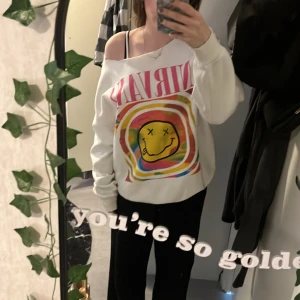 Vit Nirvana sweatshirt med färgglatt tryck - Vit sweatshirt med stort färgglatt Nirvana-tryck och smiley på framsidan. Tröjan har en lös passform och bred halsringning som kan bäras off-shoulder. Perfekt för dig som gillar bandtröjor med retrostil.
