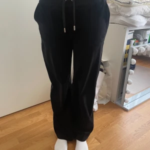 Juicy byxor - Snygga svarta byxor med snörning i midjan och fickor med knappar. Perfekta för en avslappnad stil. Byxorna har en rak passform och är tillverkade i ett mjukt material.