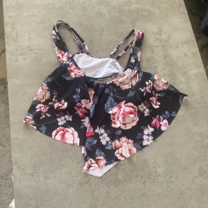 Blommig bikiniöverdel - Snygg bikiniöverdel med blommigt mönster i rosa och vita nyanser på en mörk bakgrund. Den har justerbara axelband och en volangdetalj som ger en extra touch. Perfekt för sommarens stranddagar!