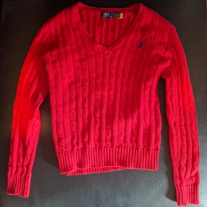 Röd stickad tröja från Polo Ralph Lauren - Säljer en klassisk röd stickad tröja från Polo Ralph Lauren med v-ringning och kabelstickat mönster. Tröjan har långa ärmar och en liten blå broderad logga på bröstet. Perfekt för lager-på-lager och snygg till jeans.