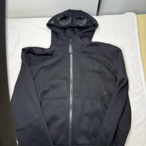 Svart hoodie med glasögon från C.P. Company - Säljer en svart hoodie från C.P. Company med dragkedja och unika inbyggda glasögon i huvan. Jackan har två fickor framtill och diskret logga på bröstet. Perfekt för dig som vill sticka ut med en streetwear-look.