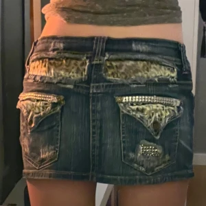 Mörk jeanskjol med leoparddetaljer från Bershka - Säljer en mörkblå jeanskjol från Bershka med snygga leopardmönstrade detaljer på bakfickorna och slitning framtill. Kjolen har pärlprydda detaljer vid fickorna och klassisk femficksdesign. Perfekt för dig som gillar en edgy och trendig stil.