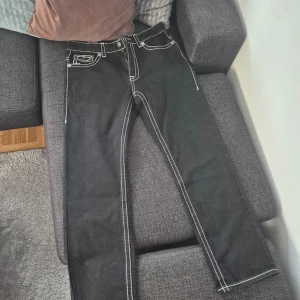 Svarta jeansbyxor från True Religion med vit kontrastsöm - Svarta jeansbyxor från True Religion med tydliga vita kontrastsömmar och klassiska bakfickor med broderad design. Byxorna har normal passform och raka ben. Märkespatch baktill i midjan och detaljerade fickor fram och bak.