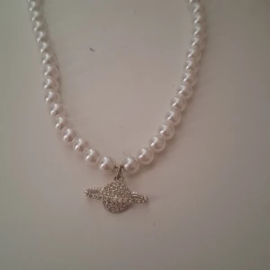 Pärlhalsband med hängsmycke - Elegant pärlhalsband med ett glittrande hängsmycke. Pärlorna är vita och hängsmycket är dekorerat med små stenar som ger en lyxig känsla. Perfekt för att lyfta vilken outfit som helst.