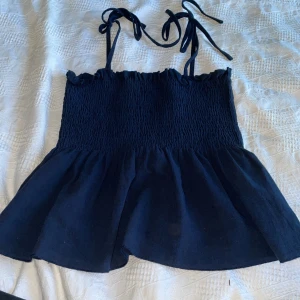 Svart ärmlös topp med knytband - Säljer en söt svart ärmlös topp med knytband på axlarna. Toppen har en smockad överdel och en luftig nederdel som ger en fin siluett. Perfekt för varma dagar!