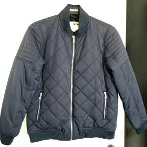 Mörkblå quiltad bomberjacka från Junkyard - Snygg mörkblå bomberjacka från Junkyard med quiltat mönster och dragkedja framtill. Jackan har långa ärmar och ribbade muddar vid ärmslut och nederkant. Perfekt för en stilren look.