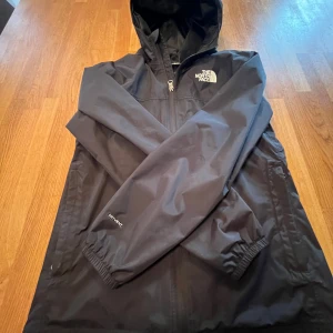 Svart vindjacka från The North Face - Säljer en svart vindjacka från The North Face med huva och dragkedja. Perfekt för blåsiga dagar och håller dig skyddad mot väder och vind. Nästan aldrig använd! 