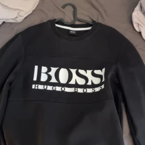 Svart tröja från Hugo Boss - Snygg svart tröja från Hugo Boss med stort vitt logotryck på framsidan. Tröjan har en klassisk rund halsringning och långa ärmar. Perfekt för en stilren och avslappnad look.