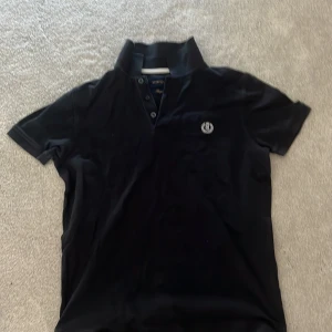 Svart pikétröja från Henri Lloyd - Snygg svart pikétröja från Henri Lloyd med korta ärmar och klassisk krage. Tröjan har knappar framtill och ett diskret broderat emblem på bröstet. Perfekt för en stilren look. Den är äkta 