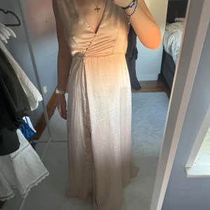 Balklänning - Champagnefärgad/ljusrosa långklänning med omlott-design och djup v-ringning. Klänningen har en slits framtill och en öppen rygg med knytband. Perfekt för speciella tillfällen.