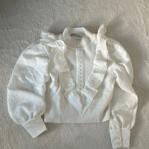 Vit blus med volanger och knappar - Elegant vit blus med volanger och dekorativa knappar framtill. Blusen har långa ärmar med puffiga axlar och ribbad krage. Perfekt för en stilfull look.