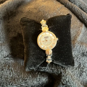 Versus Versace damklocka med gulddetaljer - Säljer en snygg klocka från Versus Versace med rund urtavla, guldfärgade detaljer och glittriga stenar runt urtavlan. Armbandet är i silverfärgat metall med guldiga emblem. Perfekt accessoar för att lyfta din stil.