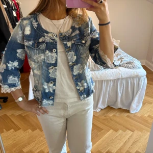 Blommig jeansjacka från Fracomina - Säljer en blå jeansjacka från Fracomina med vita blommiga mönster och trekvartsärmar med spetsdetaljer. Jackan har rund halsringning och knappar framtill.