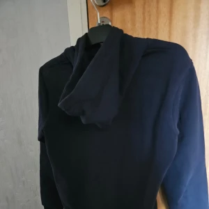 Mörkblå hoodie från GANT - Säljer en stilren mörkblå hoodie från GANT med broderad logga på bröstet. Tröjan har en klassisk huva med dragsko och ribbade muddar vid ärmslut och nederkant. Perfekt för en avslappnad stil. Svårt att fånga rätt färg på bild. Den är i värdigt i ny skick 