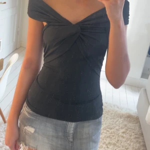 Svart offshoulder topp med twist framtill - Snygg svart topp med offshoulder-design och twistad detalj framtill. Toppens passform är tight och den har korta ärmar som sitter nedanför axlarna. Perfekt för en trendig och stilren look.