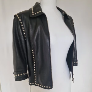 Dolce & Gabbana Vintage 2000-tal läderjacka  - Sällsynt och eftertraktad Dolce & Gabbana vintage läderjacka från tidigt 2000-tal. Tillverkad i exklusivt, mjukt läder med ett lyxigt silkesfoder,dekorerad med silverfärgade nitar.Jackan är i fint vintage skick. Lädret är mjukt och välbevarat. Små tecken på användning, inget som påverkar helhetsintrycket. Det finns en knappt synlig fläck framtill, kan nog tvättas bort.(bild) Fodret av silke med lite slitage på insidan. Skicka gärna ett meddelande för mer info och bilder 🫶