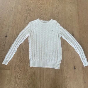 Vit kabelstickad tröja från GANT - Säljer en klassisk vit kabelstickad tröja från GANT med rund halsringning och diskret broderad logga på bröstet. Tröjan har långa ärmar och ribbade muddar vid ärmslut och nederkant. Perfekt för en stilren och tidlös look. Bra skick och en bra tröja för sena sommarkvällar😁.               Pris kan diskuteras.