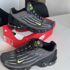 Nike Air Max Plus III Carbon Grey/Black - Nike Air Max Plus III i carbon grey och svart med detaljer i fluorescerande grönt. Skorna har en sportig design med synliga Air Max-enheter i sulan och tunna linjer längs ovandelen. Liten Swoosh-logga i grönt på sidan och TN-detaljer vid hälen.
