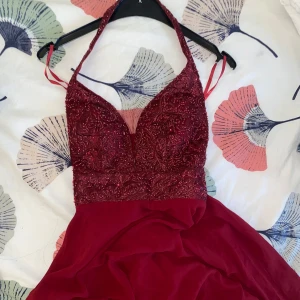 Vinröd långklänning med spets och öppen rygg - Säljer en vinröd långklänning med glittrig spets upptill och djup v-ringning. Klänningen har smala axelband, öppen rygg med snörning och en flowy kjol i mjukt tyg. Använts en gång tidigare. Snörning vid halsen gick sönder och jag har sytt ihop själv (se sista bilden), men fungerar fortfarande som den ska 💋 Orginalpris: 1000 kr! 🫰🏽