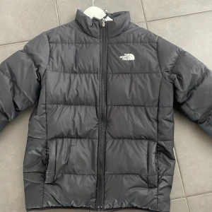 Svart pufferjacka från The North Face - Säljer en svart pufferjacka från The North Face med klassisk logga på bröstet och ryggen. Jackan har hög krage, dragkedja framtill och två sidofickor. Perfekt för kalla dagar med sin vadderade design. Går att använda på båda hållen alltså ”ut och in” finns ett litet hål men som ej är väldigt synligt. Går att laga!