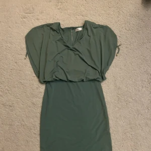 Grön kortklänning från Femme Collection - Snygg grön kortklänning från Femme Collection med loose passform och omlottliknande överdel. Klänningen har korta ärmar och ett litet nyckelhål i ryggen. Materialet är mjukt och känns som polyester, perfekt för dig som gillar stilren design med lite extra volym upptill.