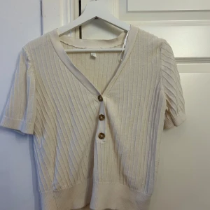 Beige ribbad blus från H&M - Kortärmad beige ribbad blus från H&M med v-ringning och tre snygga knappar framtill. Mjuk och stretchig känsla, perfekt till jeans eller kjol. Enkel och stilren design med diskreta ränder som ger en modern touch.