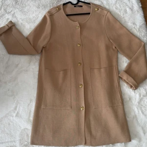 Beige cardigan jacka från Gina Tricot - Snygg beige cardigan jacka från Gina Tricot med guldknappar och stora fickor framtill. Jackan har rund halsringning, långa ärmar och axelremmar med knappdetalj. 