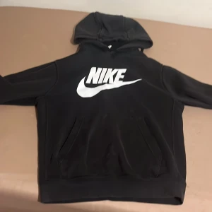 Svart Nike hoodie med stor logga - Svart hoodie från Nike med stor vit logga och swoosh på bröstet. Tröjan har huva och en klassisk känguruficka framtill. Perfekt för chill eller träning, med mjukt material och lång ärm. Snygg och enkel stil som funkar året runt.