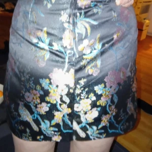 Svart blommig omlottkjol från Orsay - Snygg svart kortkjol med blommigt mönster i lila, blått och gult. Kjolen har omlott-design och ett brett bälte med metallring i midjan. Materialet är glansigt och känns lyxigt, perfekt för dig som gillar detaljer och vill sticka ut.