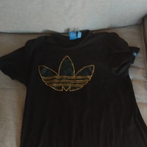 Svart Adidas t-shirt med gul logga - Svart t-shirt från Adidas med den klassiska trefoil-loggan i gult på bröstet. Skön passform och tillverkad i mjuk bomull. Perfekt för dig som gillar sportig och stilren streetwear.