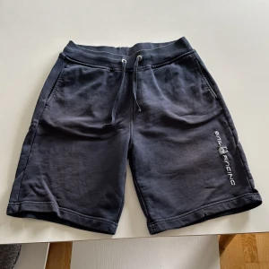 Mörkblå Sail Racing shorts med snörning - Mörkblå shorts från Sail Racing med bekväm midja och snörning framtill. Mjukt bomullsmaterial, två sidofickor och en bakficka. Snyggt Sail Racing-tryck längs benet. Perfekt för chill dagar och sportiga aktiviteter.