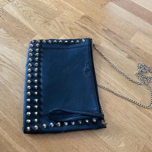 Svart kuvertväska med nitar och kedja - Cool svart kuvertväska i skinnimitation med silverfärgade nitar längs kanterna och en lång kedjerem. Väskan har dragkedja upptill och en liten handledsrem. Perfekt för dig som gillar edgy detaljer och vill sticka ut.