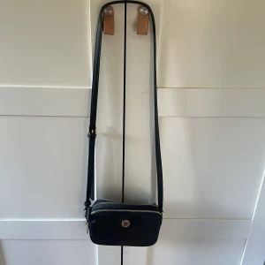 Svart axelväska från Tommy Hilfiger - Snygg svart axelväska från Tommy Hilfiger med guldiga detaljer och dubbla dragkedjor då den har dubbla fack . Väskan har justerbar rem och klassisk TH-logga framtill. 