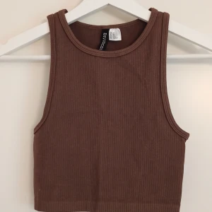 Brun ribbad croppad linnetopp H&M Divided - Brun croppad topp från H&M i storlek XS. Toppen är ribbad och har en ärmlös design med hög hals. Materialet är mjukt och stretchigt, tillverkat av 94% polyamid och 6% elastan. Inga tecken på användning. Perfekt för dig som gillar enkla och stilrena plagg.