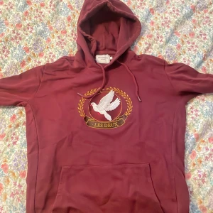 Vinröd hoodie från Les Deux med brodyr - Säljer en vinröd hoodie från Les Deux med broderad duva och gulddetaljer på bröstet. Hoodien är enbart använd ett fåtal gånger, så skicket är åtminstone 9/10