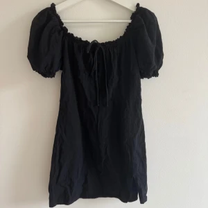 Svart kort klänning  - Supersöt svart klänning med puffärmar och smockad kant vid halsen. Klänningen har snörning framtill, en liten slits nedtill och dragkedja i ryggen. Materialet är bomull och modellen är kort, perfekt för dig som gillar en feminin och trendig look.