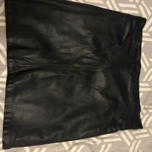 Svart skinnkjol från The Black Label - Säljer en svart skinnkjol från The Black Label Collection i storlek M. Kjolen är kort och har en stilren, rak form med diskret söm framtill. Perfekt för dig som gillar en edgy och cool look. Materialet är mjukt och följsamt skinn.