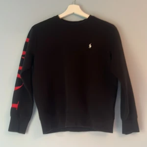 Svart sweatshirt från Polo Ralph Lauren - Svart sweatshirt från Polo Ralph Lauren i storlek M. Tröjan har lång ärm med stor röd POLO-text längs ena ärmen och den klassiska broderade loggan på bröstet. Mjuk bomullskvalitet och ribbade muddar vid ärmslut och nederkant. Perfekt för en chill och stilren look. Väldigt bra skick