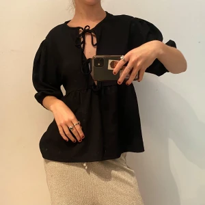 Svart blus med snörning och puffärm - Svart blus med puffiga trekvartsärmar och snygg snörning framtill vid halsen. Modellen är lite loose och har en lätt v-ringning. Perfekt för dig som gillar en chill men ändå trendig stil.