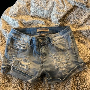 Blå jeansshorts med slitningar M - Snygga blå jeansshorts från Monday Premium i storlek M. De har coola slitningar och pärldetaljer framtill, klassisk femficksdesign och uppvikta ben. Perfekta för dig som gillar en avslappnad och trendig stil.